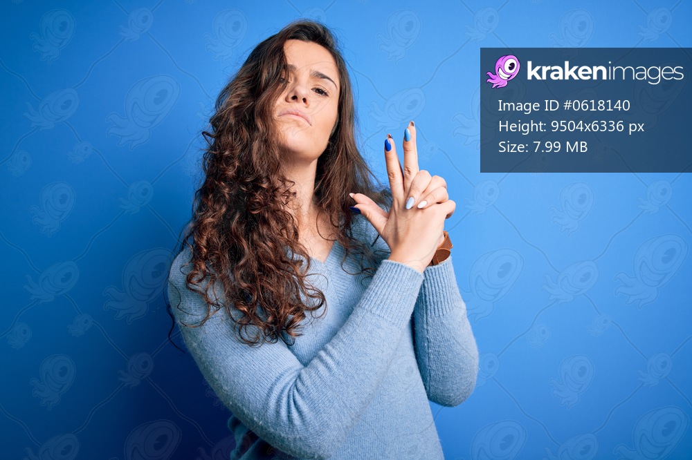 Young beautiful woman with curly hair wearing blue casual sweater over isolated background Holding symbolic gun with hand gesture, playing killing shooting weapons, angry face