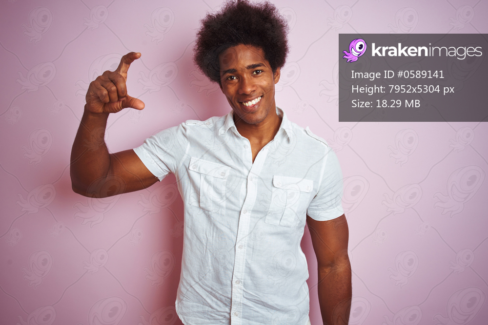 Young american man with afro hair wearing white shirt standing over isolated pink background smiling and confident gesturing with hand doing small size sign with fingers looking and the camera. Measure concept.