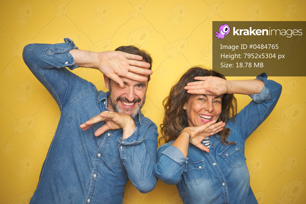 Beautiful middle age couple together standing over isolated yellow background Smiling cheerful playing peek a boo with hands showing face. Surprised and exited