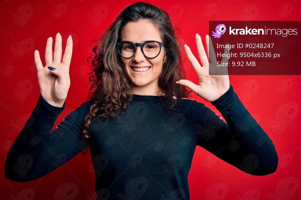 Young beautiful woman with curly hair wearing sweater and glasses over red background showing and pointing up with fingers number nine while smiling confident and happy.