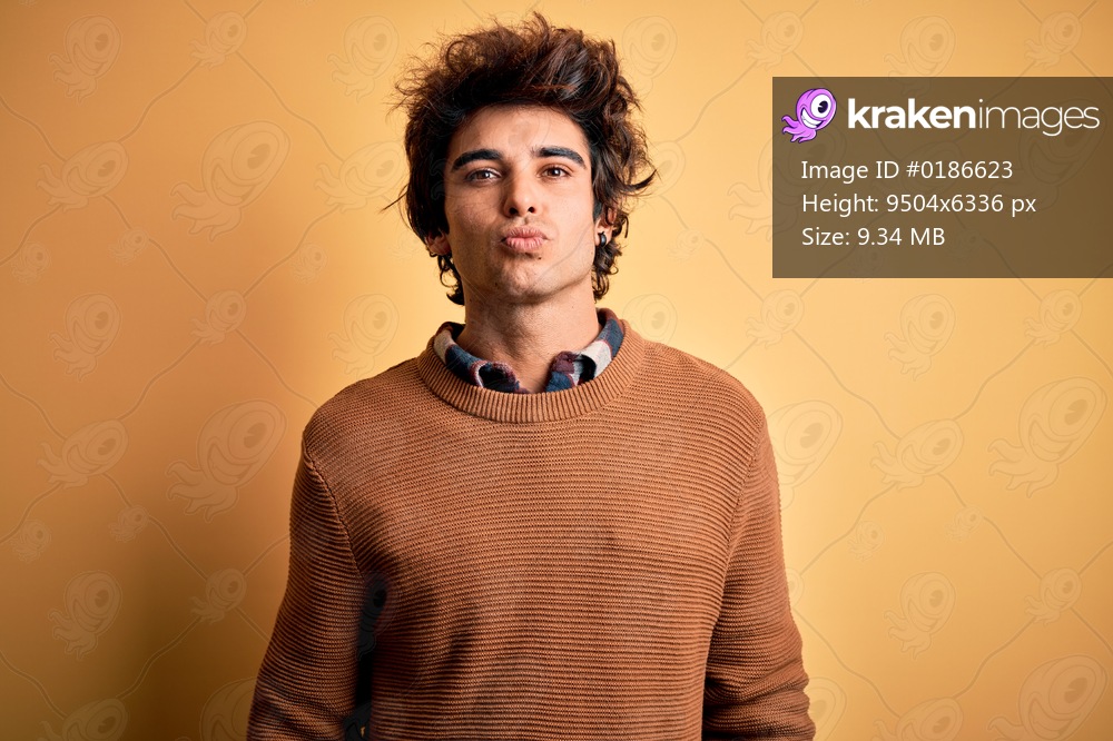 Young handsome man wearing casual shirt and sweater over isolated yellow background looking at the camera blowing a kiss on air being lovely and sexy. Love expression.