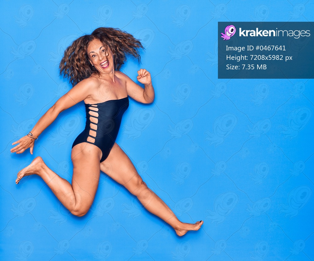 Middle age beautiful hispanic woman on vacation wearing swimsuit smiling happy. Jumping with smile on face celebrating over isolated blue background