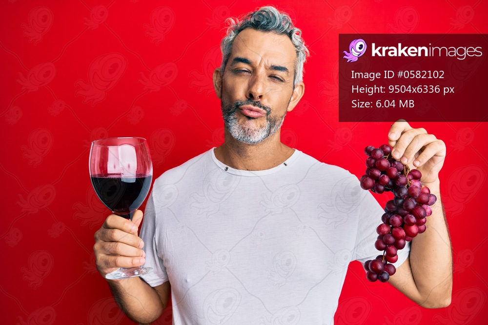 Middle age grey-haired man holding branch of fresh grapes and red wine looking at the camera blowing a kiss being lovely and sexy. love expression. 