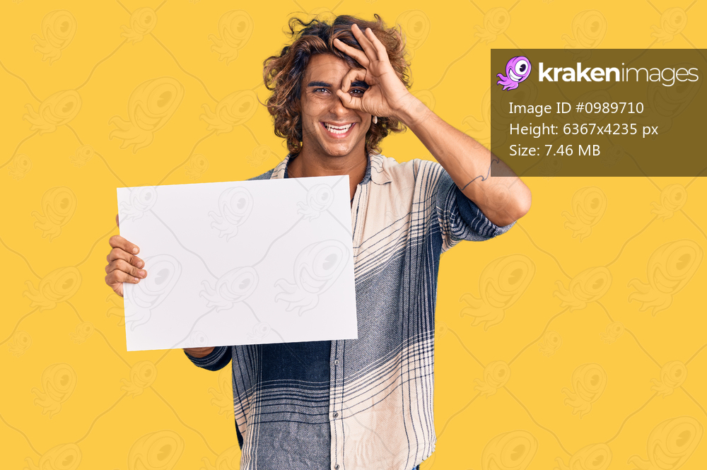Young hispanic man holding blank empty banner smiling happy doing ok sign with hand on eye looking through fingers 