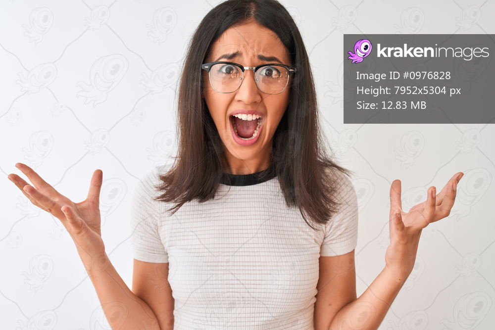Chinese woman wearing casual t-shirt and glasses standing over isolated white background crazy and mad shouting and yelling with aggressive expression and arms raised. Frustration concept.