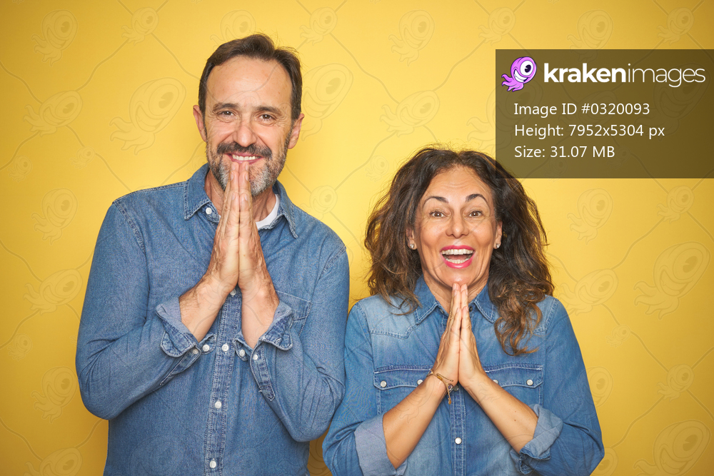 Beautiful middle age couple together standing over isolated yellow background praying with hands together asking for forgiveness smiling confident.