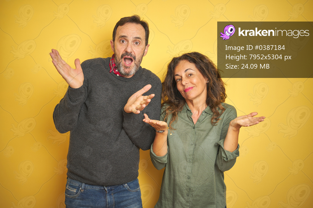 Beautiful middle age couple over isolated yellow background clueless and confused expression with arms and hands raised. Doubt concept.