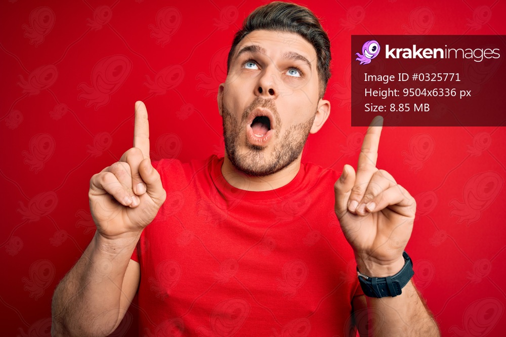 Young man with blue eyes wearing casual t-shirt over red isolated background amazed and surprised looking up and pointing with fingers and raised arms.