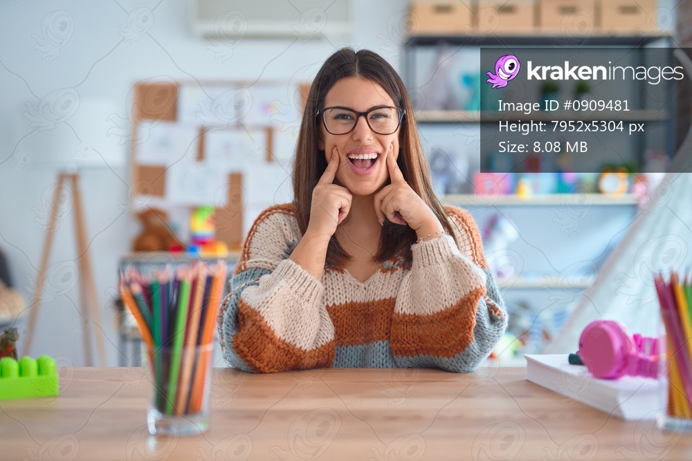 Young beautiful teacher woman wearing sweater and glasses sitting on desk at kindergarten Smiling with open mouth, fingers pointing and forcing cheerful smile