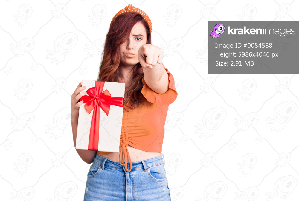 Young beautiful woman holding gift pointing with finger to the camera and to you, confident gesture looking serious 
