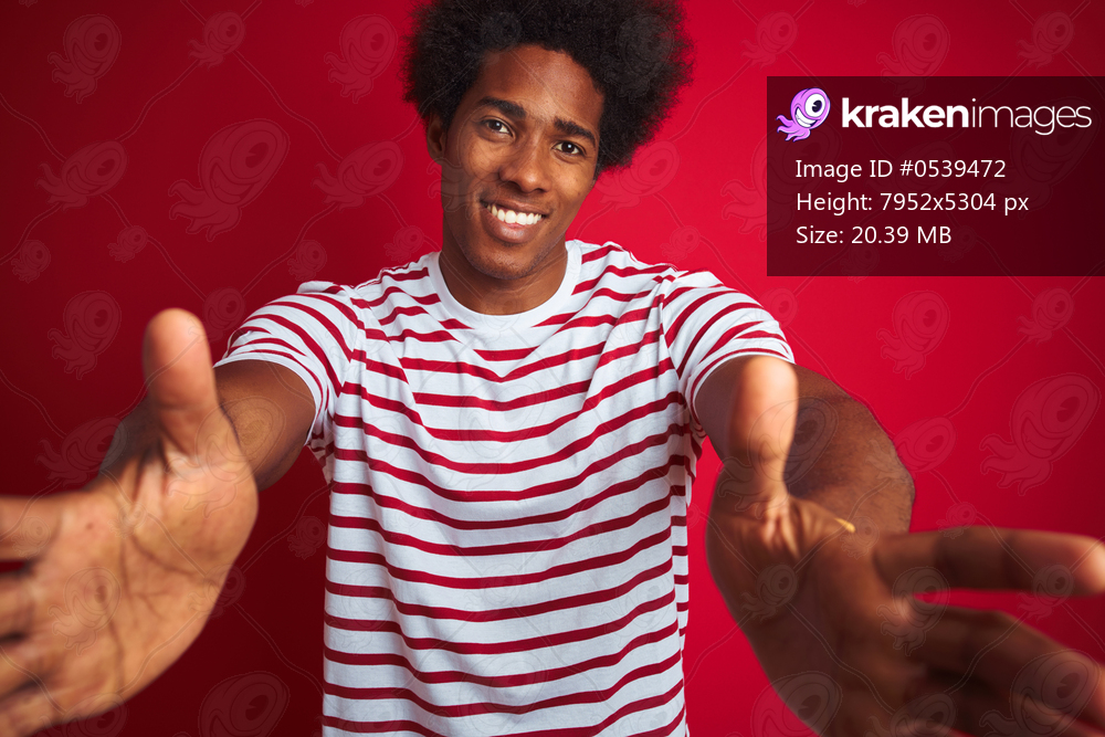 Young african american man with afro hair wearing striped t-shirt over isolated red background looking at the camera smiling with open arms for hug. Cheerful expression embracing happiness.