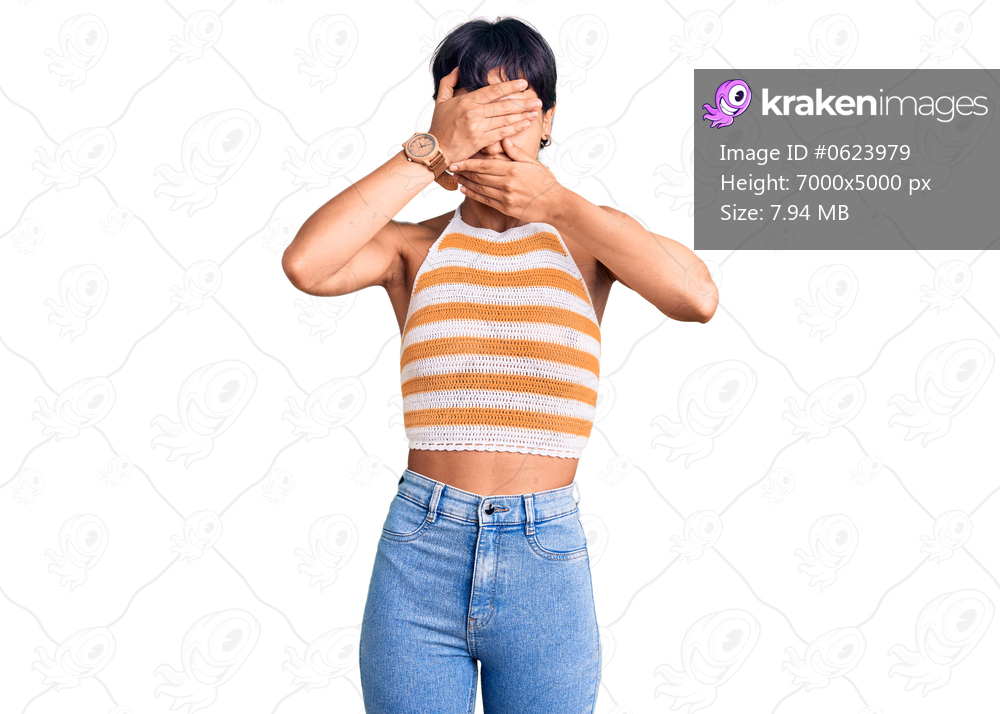 Young brunette woman with short hair wearing casual clothes covering eyes and mouth with hands, surprised and shocked. hiding emotion 