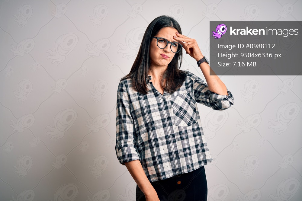 Young brunette woman with blue eyes wearing casual shirt and glasses over white background worried and stressed about a problem with hand on forehead, nervous and anxious for crisis