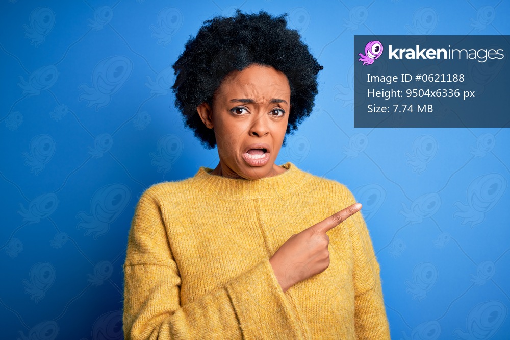 Young beautiful African American afro woman with curly hair wearing yellow casual sweater Pointing aside worried and nervous with forefinger, concerned and surprised expression