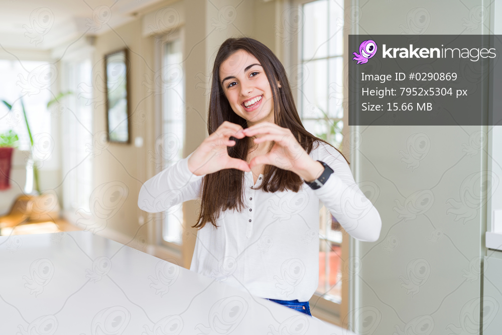 Beautiful young woman sitting on white table at home smiling in love showing heart symbol and shape with hands. Romantic concept.
