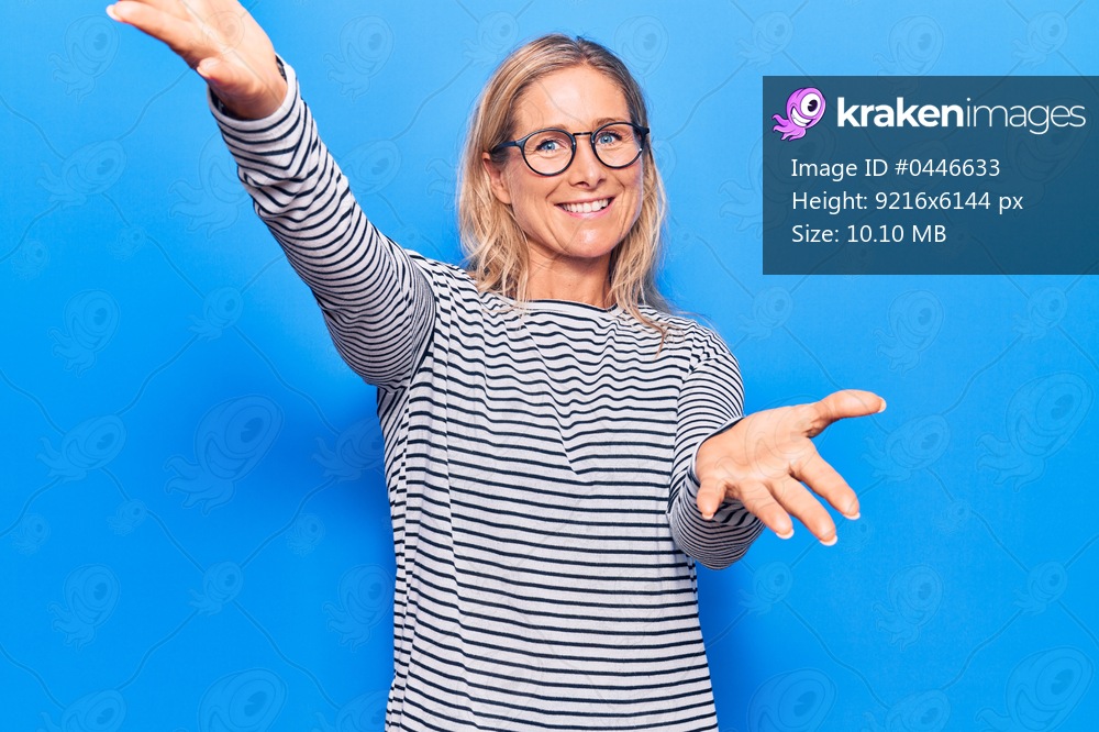 Middle age caucasian blonde woman wearing casual striped sweater and glasses looking at the camera smiling with open arms for hug. cheerful expression embracing happiness. 