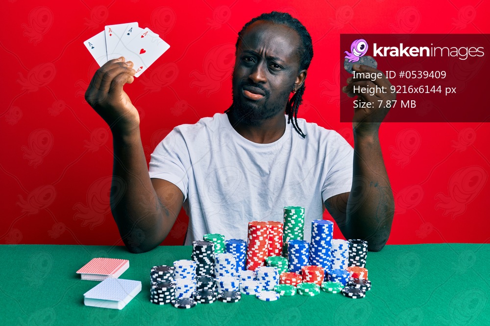 Handsome young black man sitting on the table with poker chips and cards clueless and confused expression. doubt concept. 