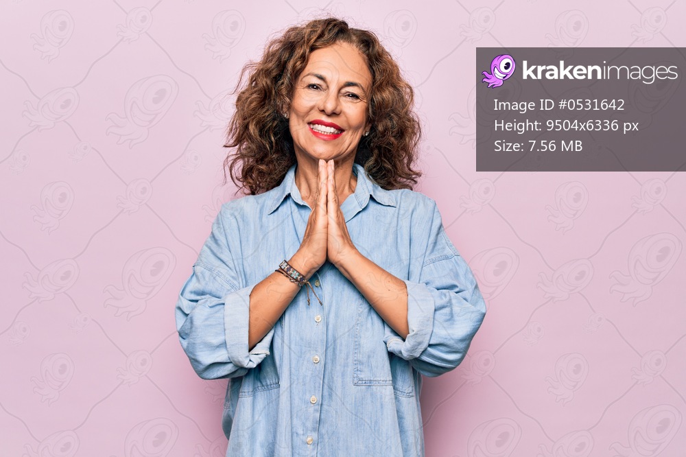 Middle age beautiful woman wearing casual denim shirt standing over pink background praying with hands together asking for forgiveness smiling confident.