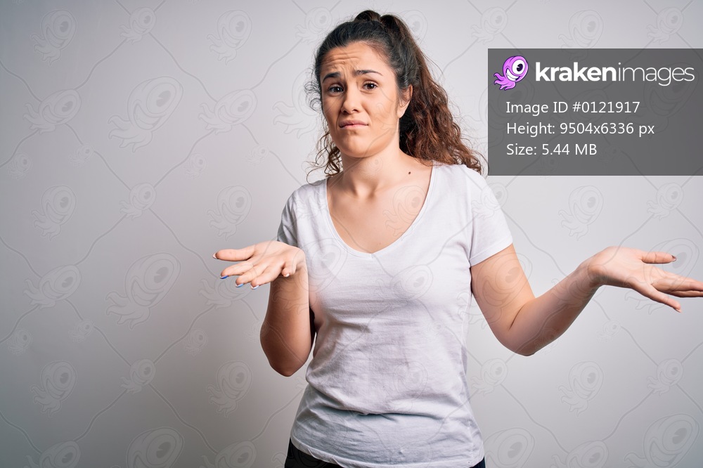 Young beautiful woman with curly hair wearing casual t-shirt standing over white background clueless and confused expression with arms and hands raised. Doubt concept.