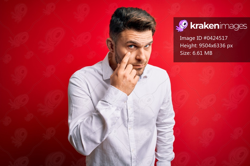 Young business man with blue eyes wearing elegant shirt standing over red isolated background Pointing to the eye watching you gesture, suspicious expression