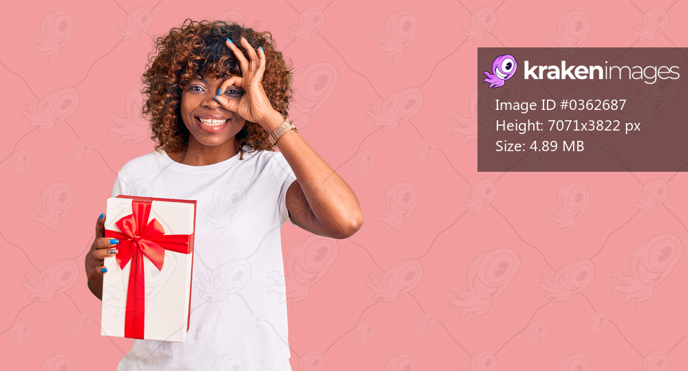 Young african american woman holding gift smiling happy doing ok sign with hand on eye looking through fingers 