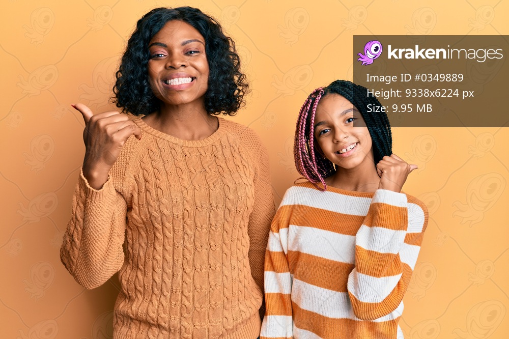 Beautiful african american mother and daughter wearing wool winter sweater pointing to the back behind with hand and thumbs up, smiling confident 