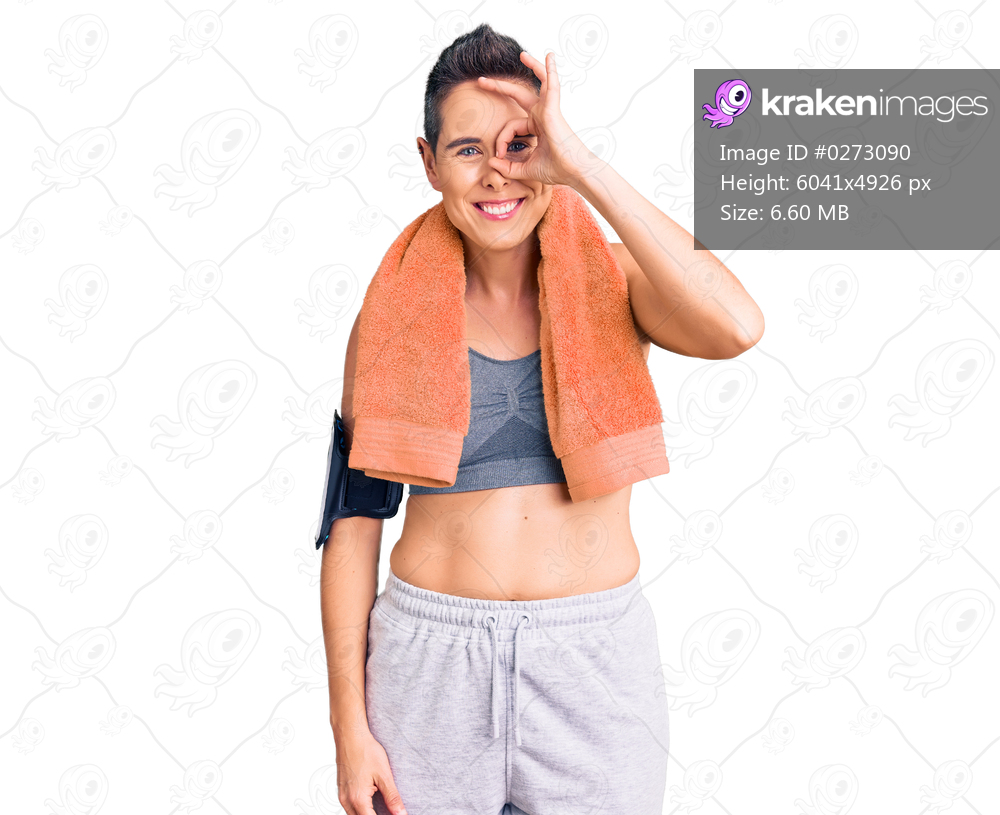 Young woman with short hair wearing sportswear and towel using smartphone doing ok gesture with hand smiling, eye looking through fingers with happy face. 