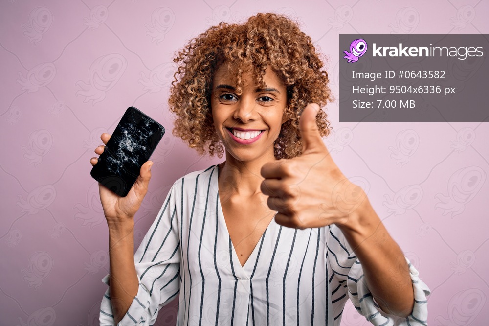Young african american curly woman holding broken smartphone showing craked screen happy with big smile doing ok sign, thumb up with fingers, excellent sign