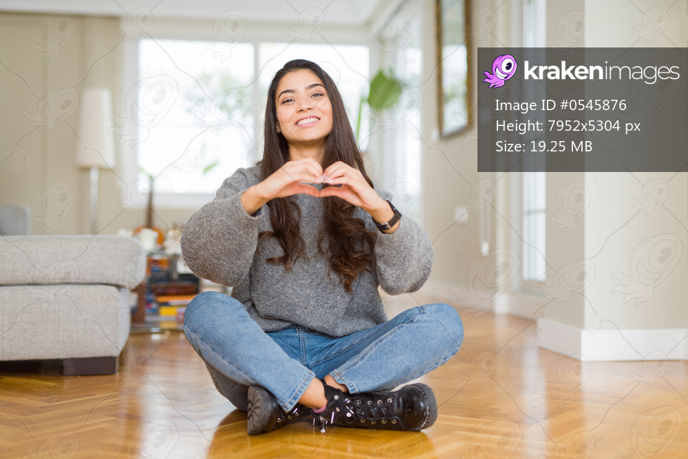 Young beautiful woman sitting on the floor at home smiling in love showing heart symbol and shape with hands. Romantic concept.