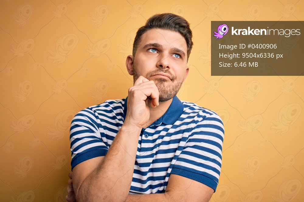 Young man with blue eyes wearing casual striped t-shirt over yellow background with hand on chin thinking about question, pensive expression. Smiling with thoughtful face. Doubt concept.