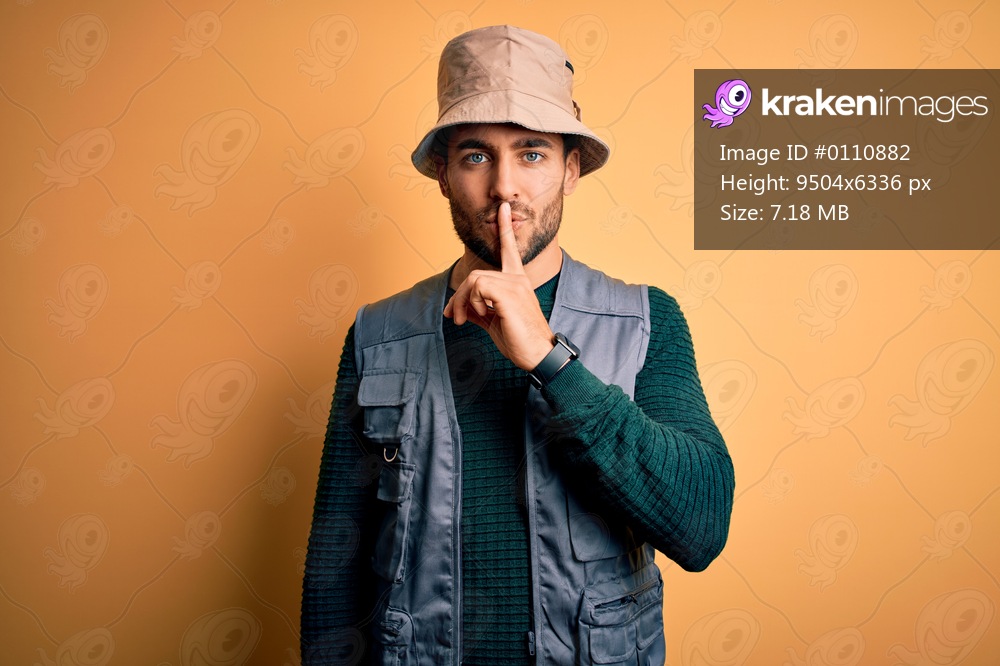 Handsome tourist man with beard on vacation wearing explorer hat over yellow background asking to be quiet with finger on lips. Silence and secret concept.