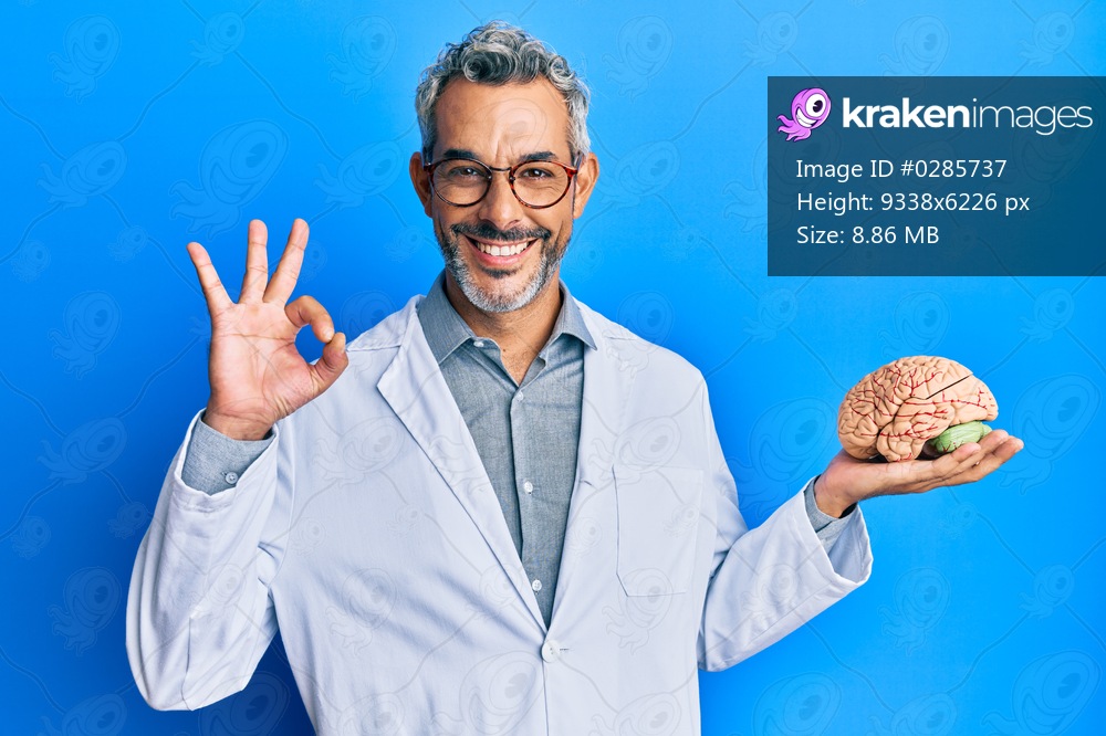 Middle age grey-haired man wearing doctor coat holding brain doing ok sign with fingers, smiling friendly gesturing excellent symbol 