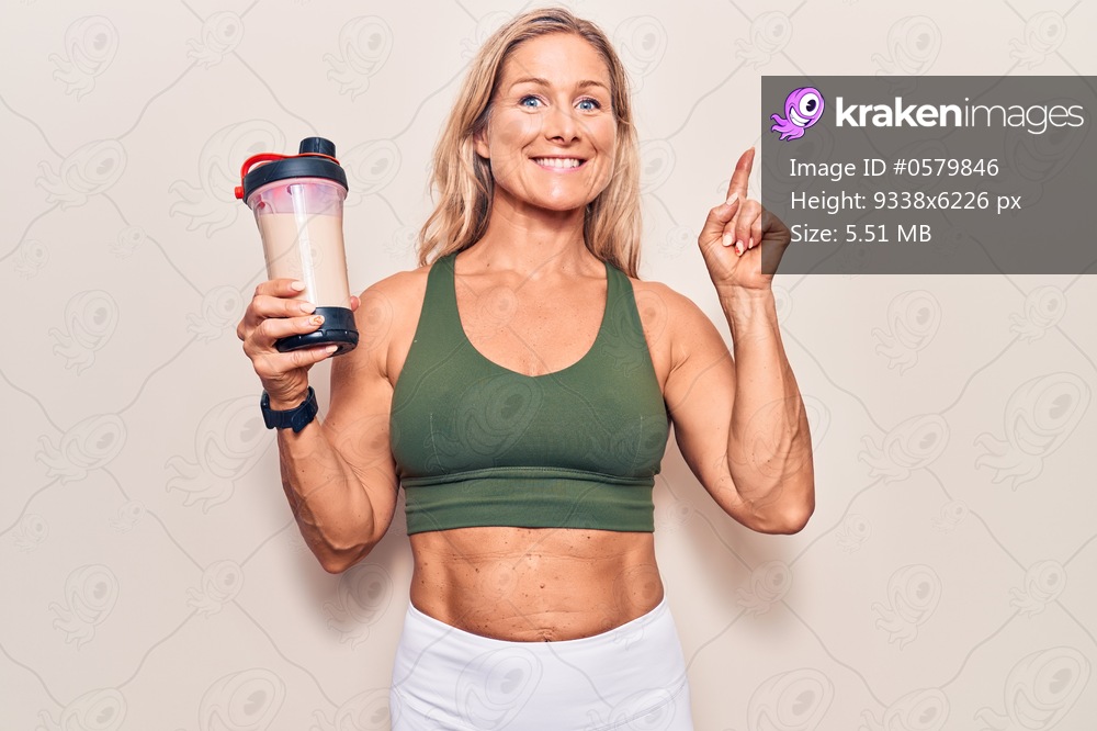 Middle age caucasian blonde woman wearing sport clothes drinking a protein shake surprised with an idea or question pointing finger with happy face, number one 