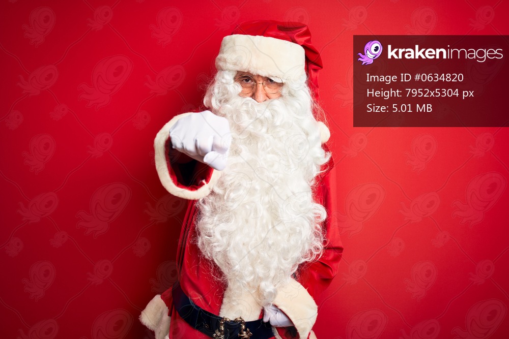 Middle age handsome man wearing Santa costume standing over isolated red background Punching fist to fight, aggressive and angry attack, threat and violence