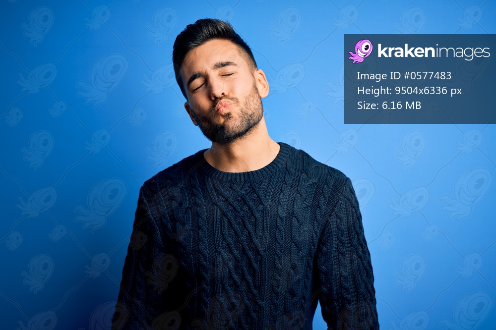 Young handsome man wearing casual sweater standing over isolated blue background looking at the camera blowing a kiss on air being lovely and sexy. Love expression.