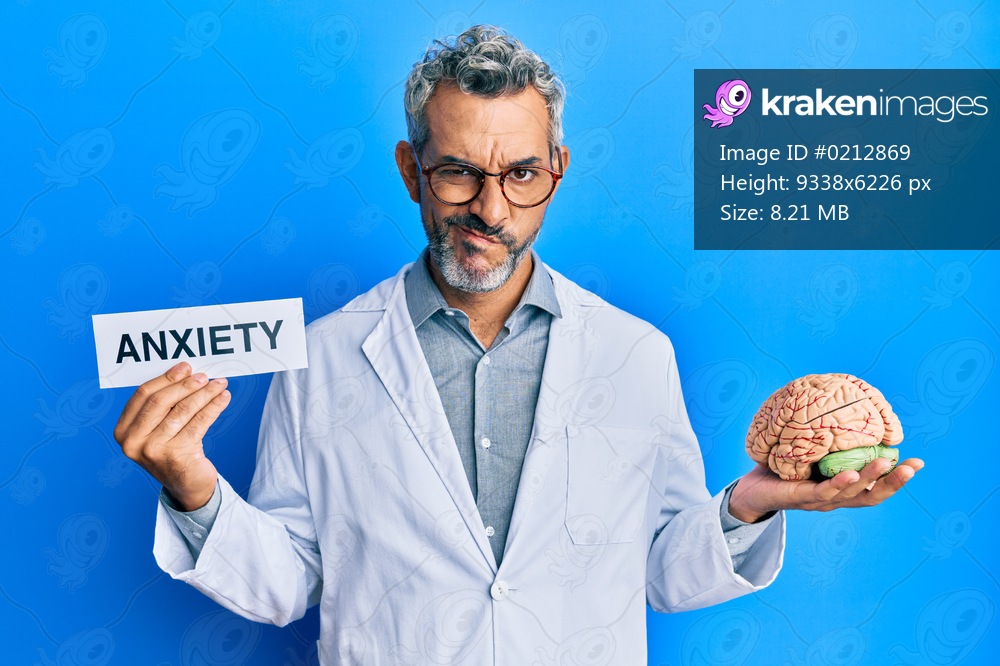 Middle age grey-haired man wearing doctor coat holding brain and anxiety message skeptic and nervous, frowning upset because of problem. negative person. 