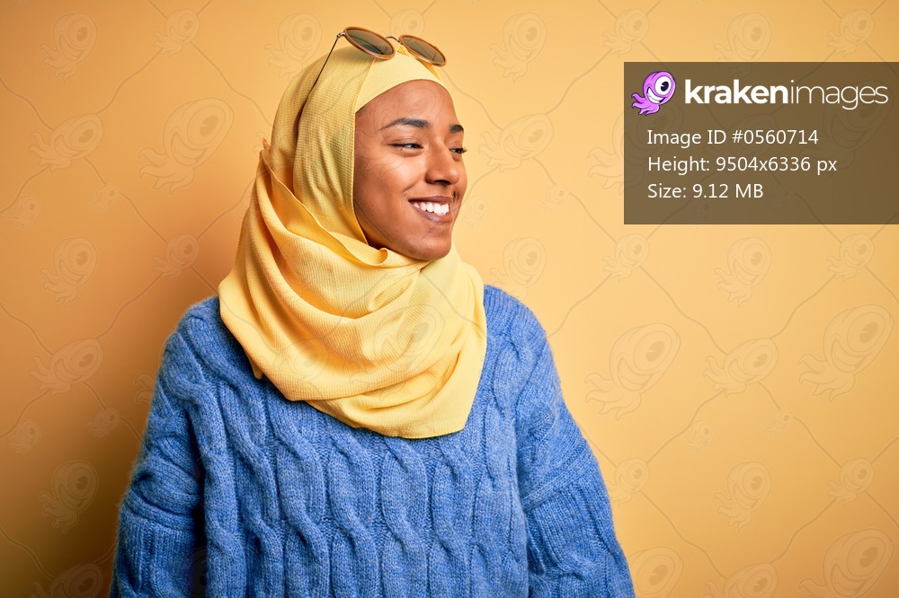 Young African American student woman wearing yellow muslim hijab and sunglasses looking away to side with smile on face, natural expression. Laughing confident.