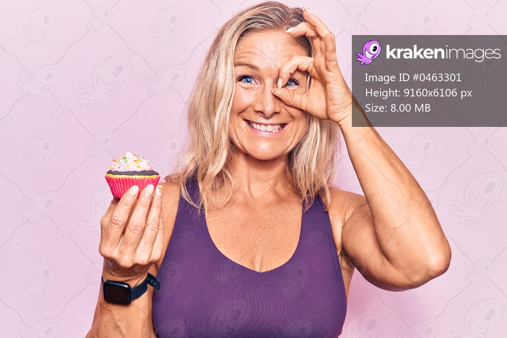 Middle age caucasian blonde woman wearing sportswear holding cupckae smiling happy doing ok sign with hand on eye looking through fingers 