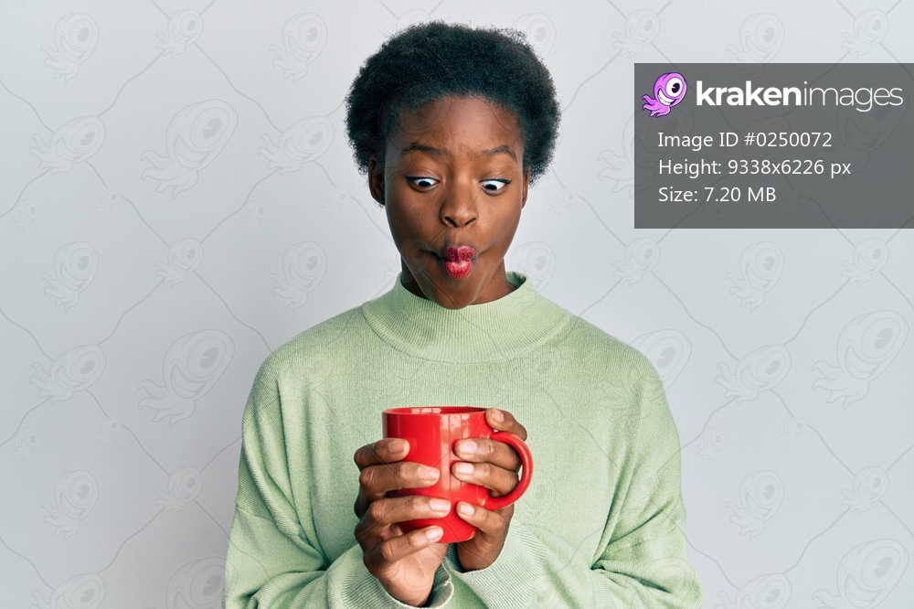 Young african american girl drinking a cup of coffee making fish face with mouth and squinting eyes, crazy and comical. 