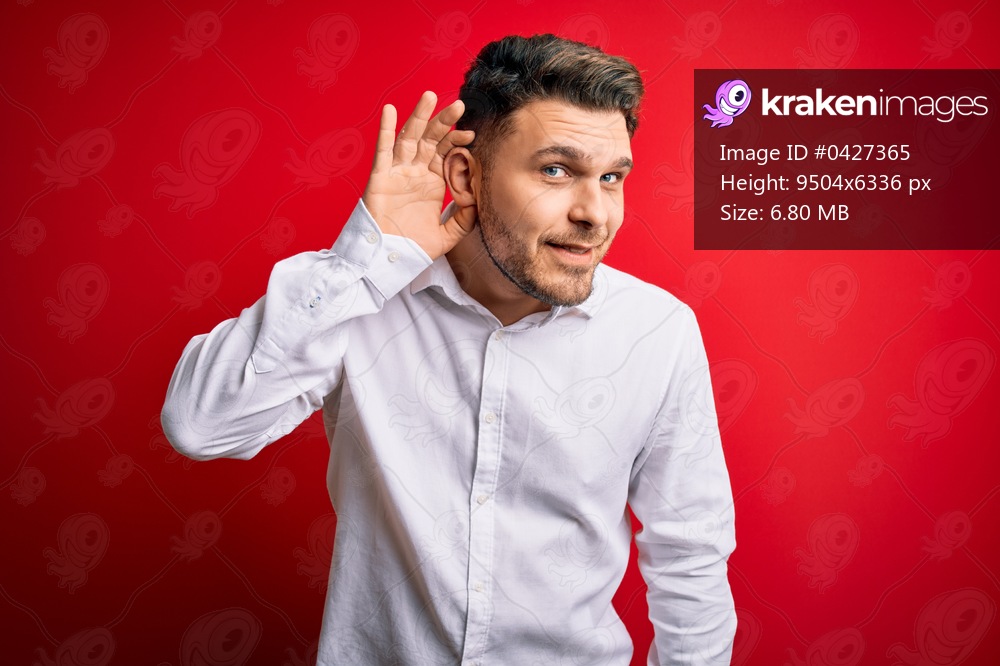 Young business man with blue eyes wearing elegant shirt standing over red isolated background smiling with hand over ear listening an hearing to rumor or gossip. Deafness concept.