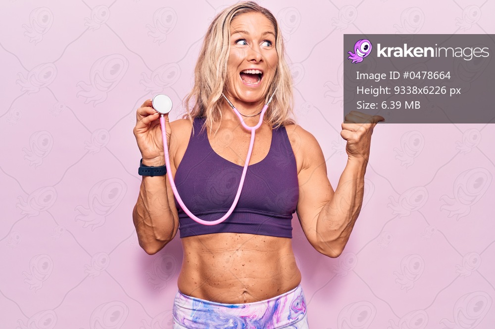 Middle age caucasian blonde woman wearing sport clothes holding stethoscope pointing thumb up to the side smiling happy with open mouth 