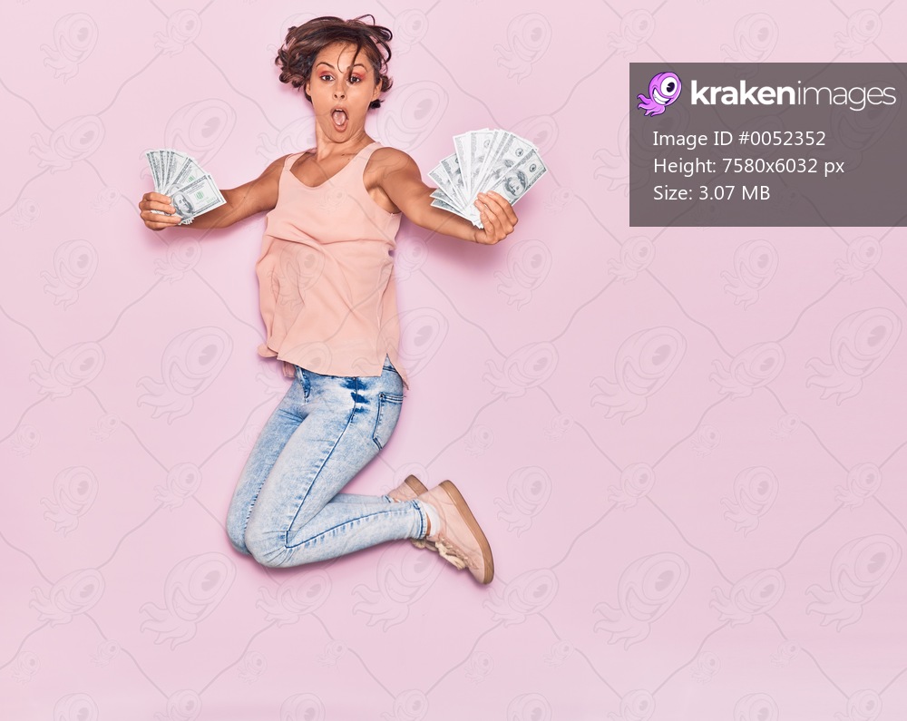 Young beautiful woman surprised holding dollars banknotes . Jumping with open mouth over isolated pink background.