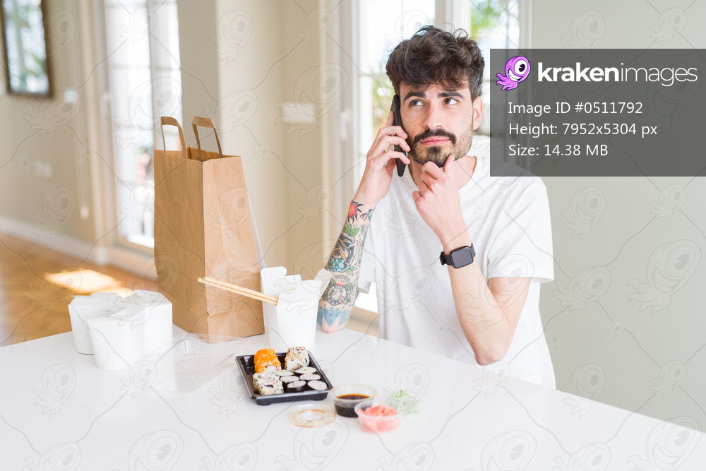 Young man eating asian sushi from home delivery and ordering food talking on smartphone serious face thinking about question, very confused idea
