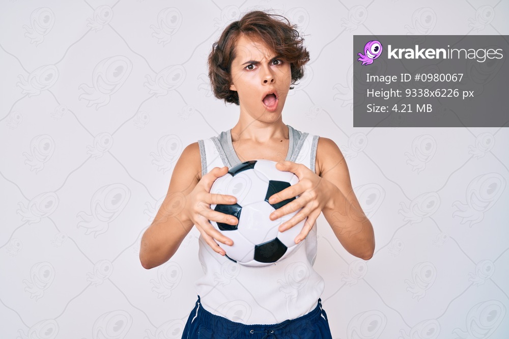Young hispanic woman holding soccer ball in shock face, looking skeptical and sarcastic, surprised with open mouth 
