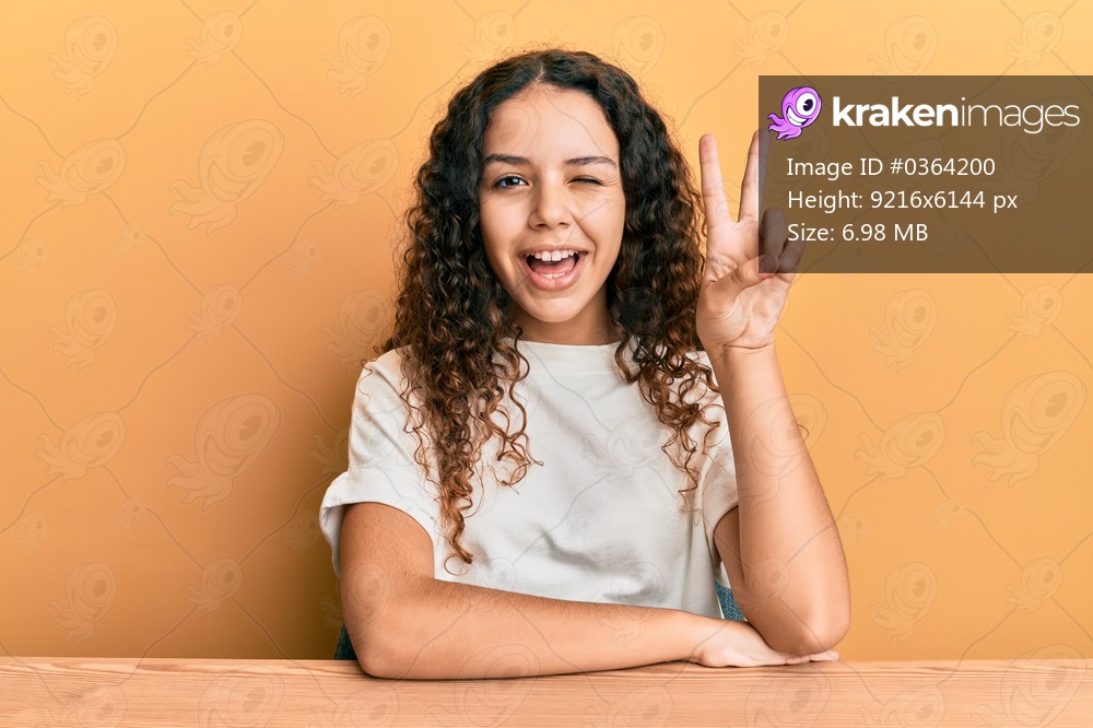 Teenager hispanic girl wearing casual clothes sitting on the table smiling with happy face winking at the camera doing victory sign. number two. 