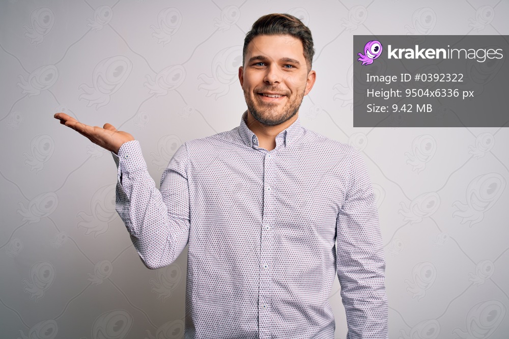 Young business man with blue eyes standing over isolated background smiling cheerful presenting and pointing with palm of hand looking at the camera.
