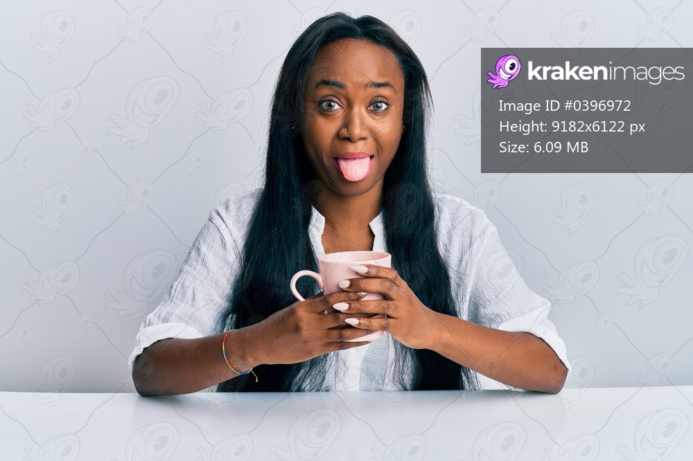 Young african woman drinking a cup coffee sticking tongue out happy with funny expression. 