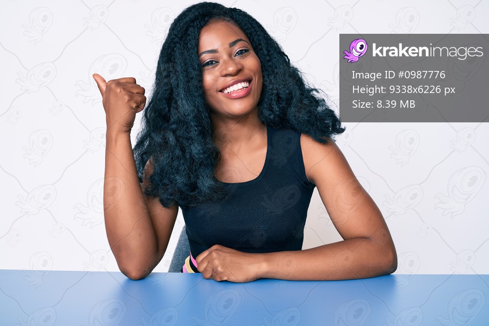 Beautiful african woman wearing casual clothes sitting on the table smiling with happy face looking and pointing to the side with thumb up. 