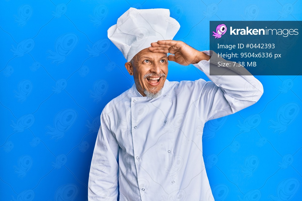 Middle age grey-haired man wearing professional cook uniform and hat very happy and smiling looking far away with hand over head. searching concept. 