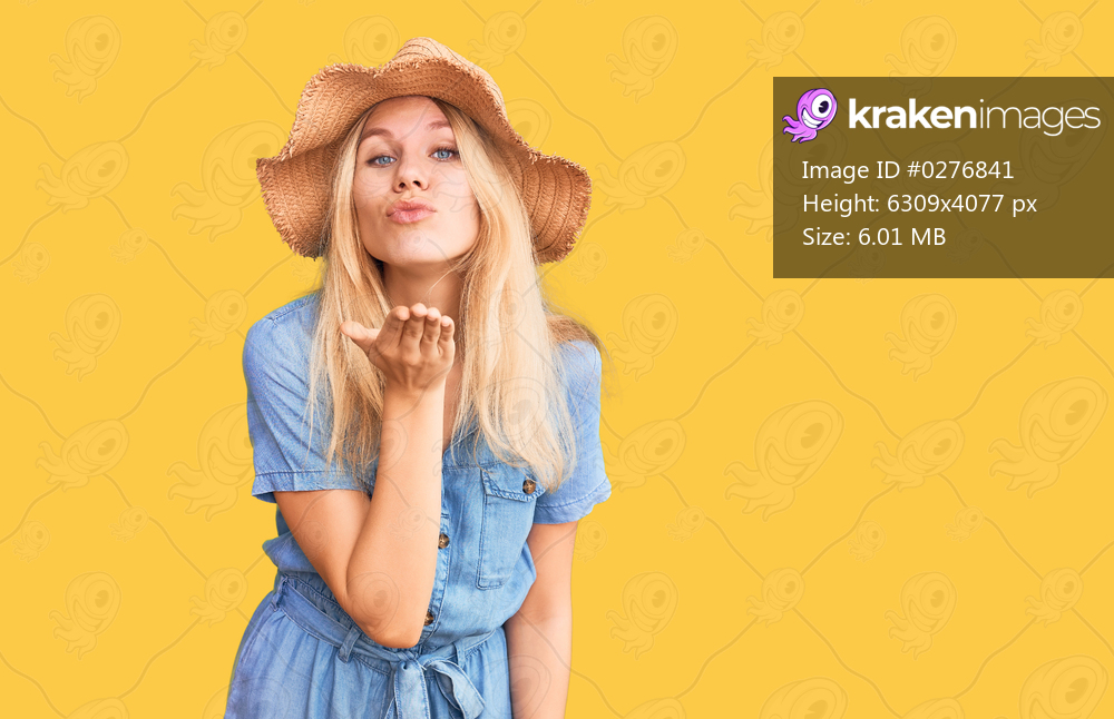 Young beautiful blonde woman wearing summer hat and dress looking at the camera blowing a kiss with hand on air being lovely and sexy. love expression. 